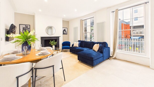 A modern living room with a dining area, a blue sofa set, and large windows with a view of buildings. - *NEW* Artisan Court | Central | Modern | Parking (Gloucestershire)