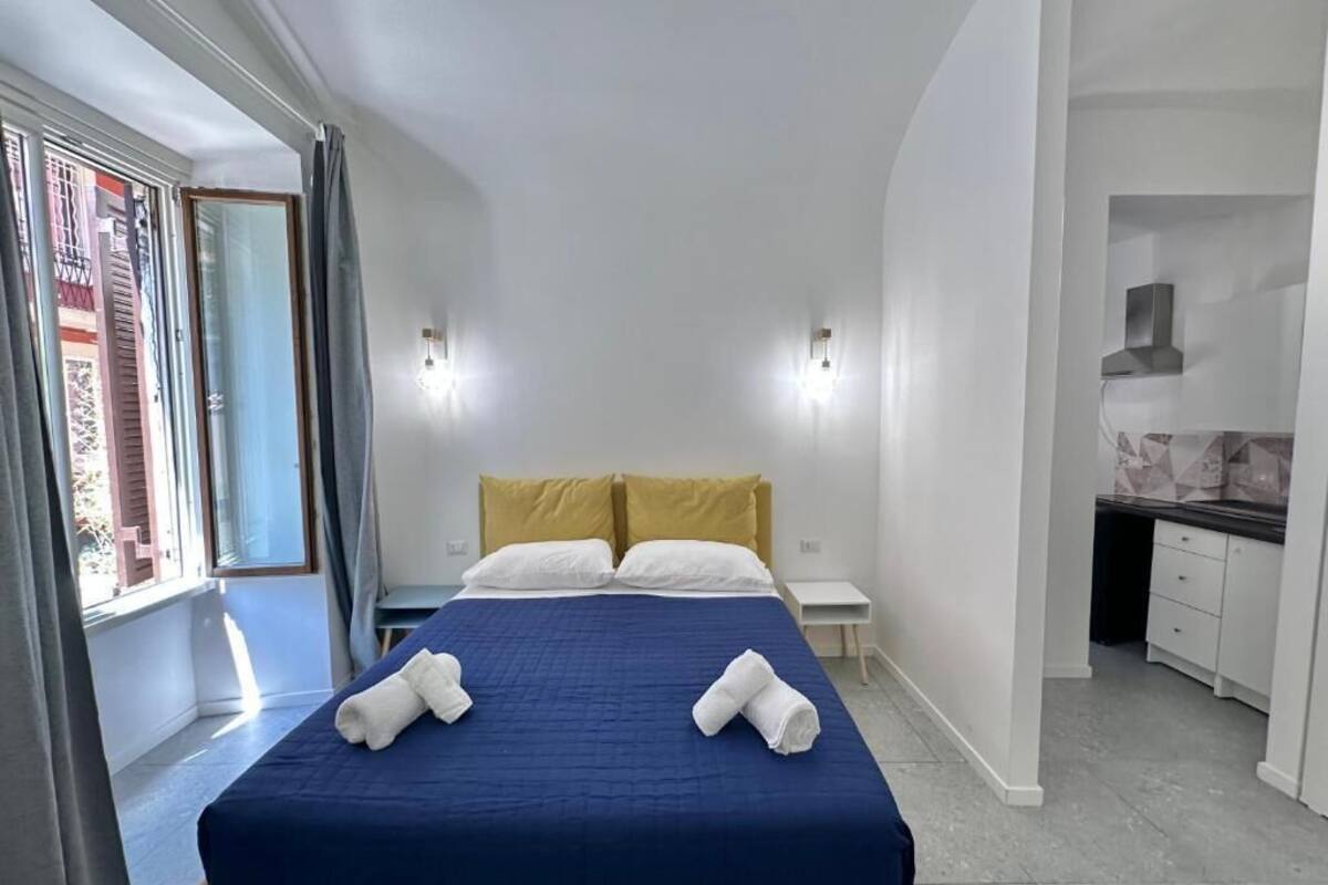 A hotel room with a bed, a small bedside table, a nightstand, and a kitchenette area.