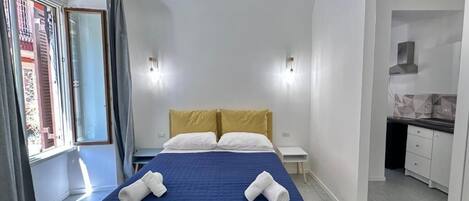 A hotel room with a bed, a small bedside table, a nightstand, and a kitchenette area.