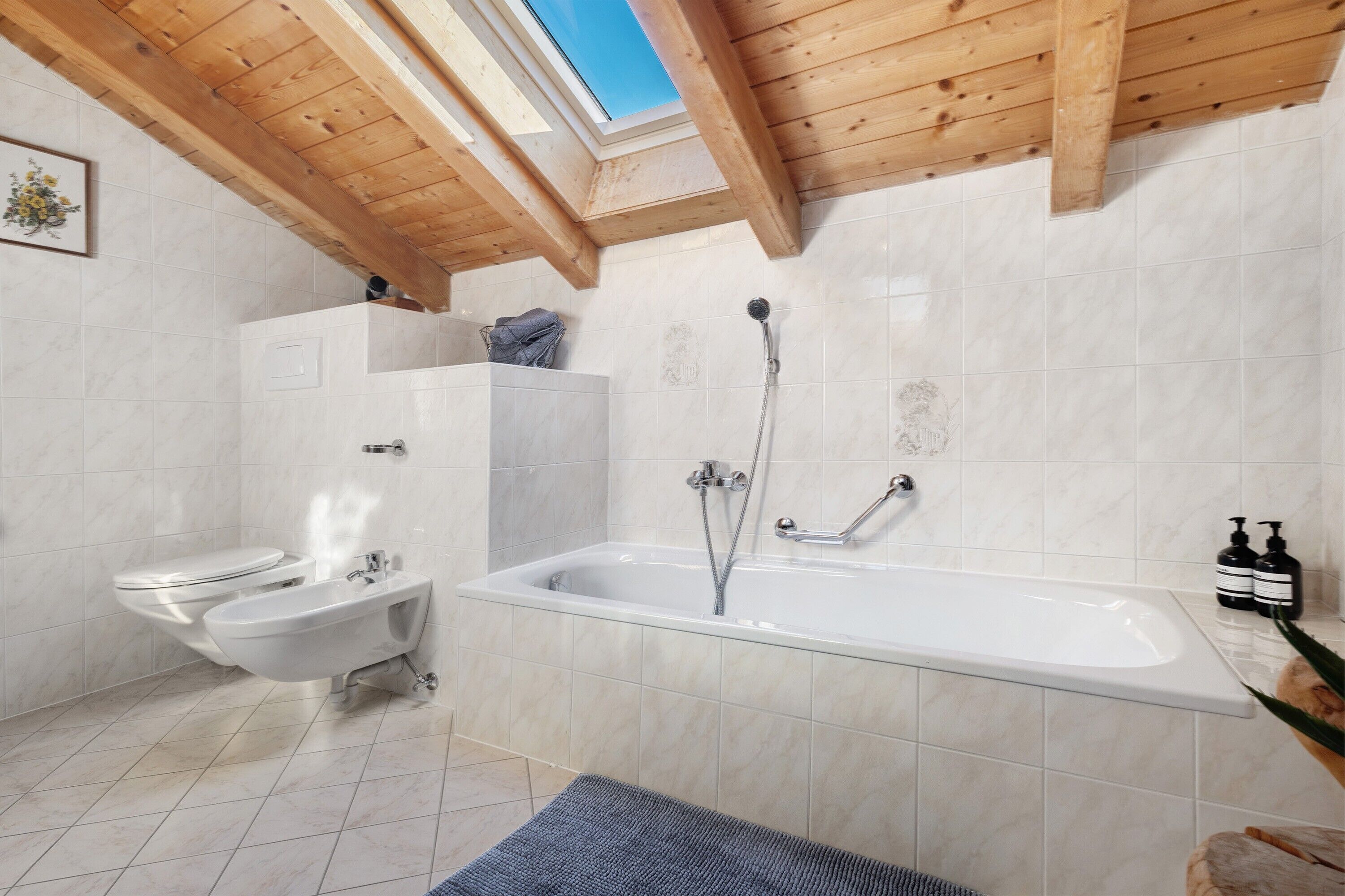 Une salle de bain moderne équipée d’un bidet, de toilettes et d’une baignoire, avec des poutres en bois au plafond et un puits de lumière.