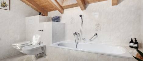 Une salle de bain moderne équipée d’un bidet, de toilettes et d’une baignoire, avec des poutres en bois au plafond et un puits de lumière.