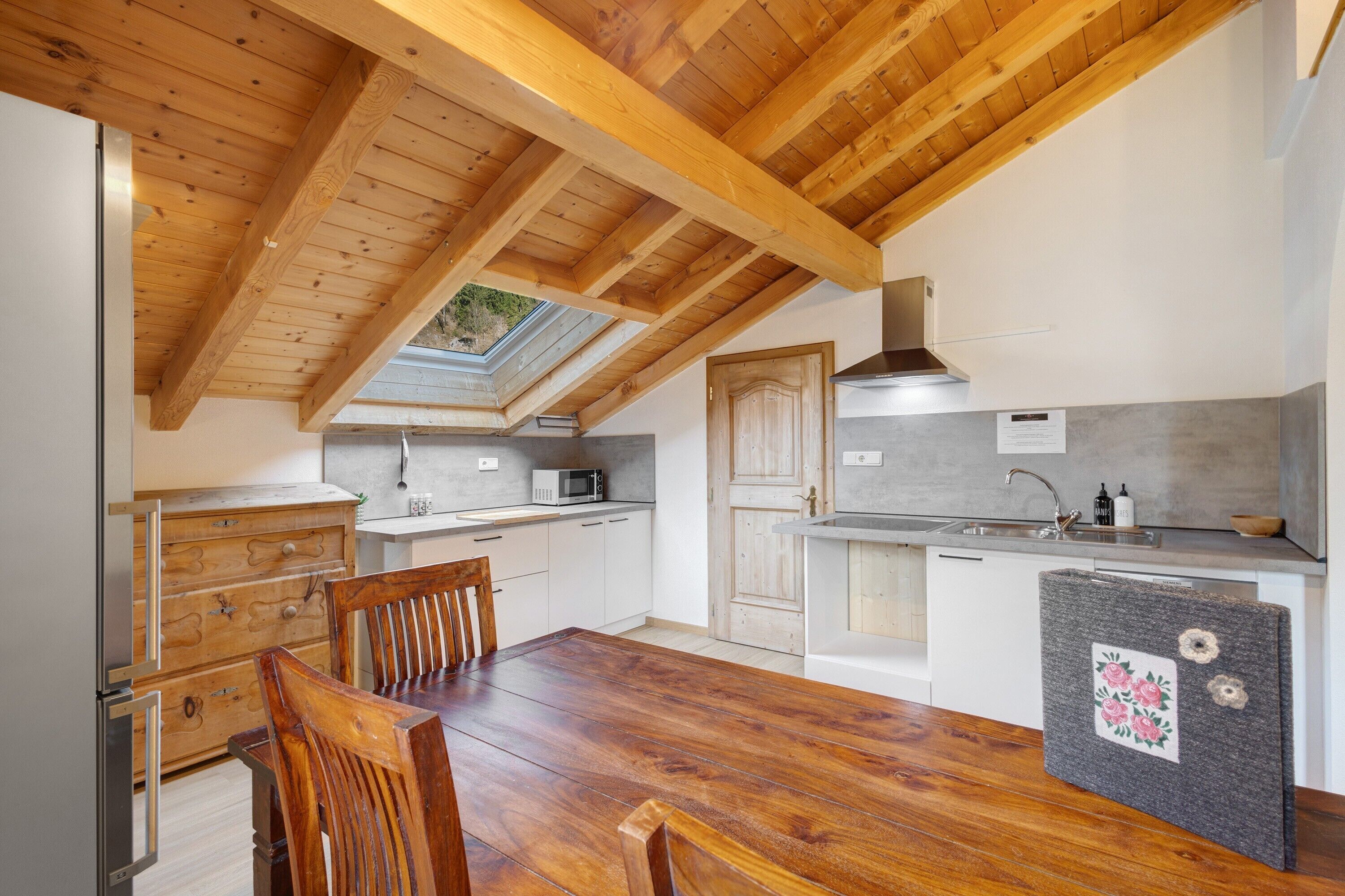 A kitchen with a wooden ceiling, a dining table, chairs, a refrigerator, a microwave, cabinets, and a skylight.