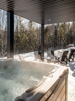 Ein Whirlpool mit Holzrand, umgeben von mit Schnee bedeckten Stühlen und Tischen, vor einer Baumlandschaft.