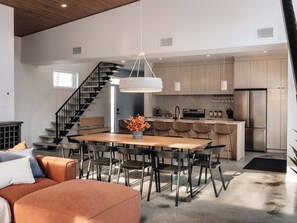 A modern kitchen with a central island, a dining area with a wooden table and chairs, and a staircase leading to an upper level. - Vitalité | Private Spa, Sauna & Pool table (Saint-Côme)