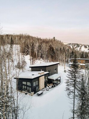 A modern two-story house with a large glass balcony, surrounded by snow-covered trees and a mountainous backdrop. - Vitalité | Private Spa, Sauna & Pool table (Saint-Côme)