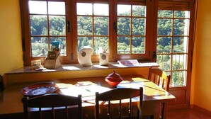 A dining area with a table and chairs, large windows with a view of a forest, and a window sill with various items. - Apartamentos Rurales Veredas (Santa Eulalia de Oscos)