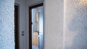 A narrow hallway leading to a kitchen with a wooden door and a light switch on the wall. - Casa Eleonora (Zogno)