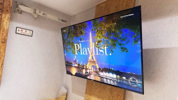 A flat-screen TV mounted on a wall displaying a 'Playlist' with the Eiffel Tower in the background. - ORAGO stay hotel (Suwon)