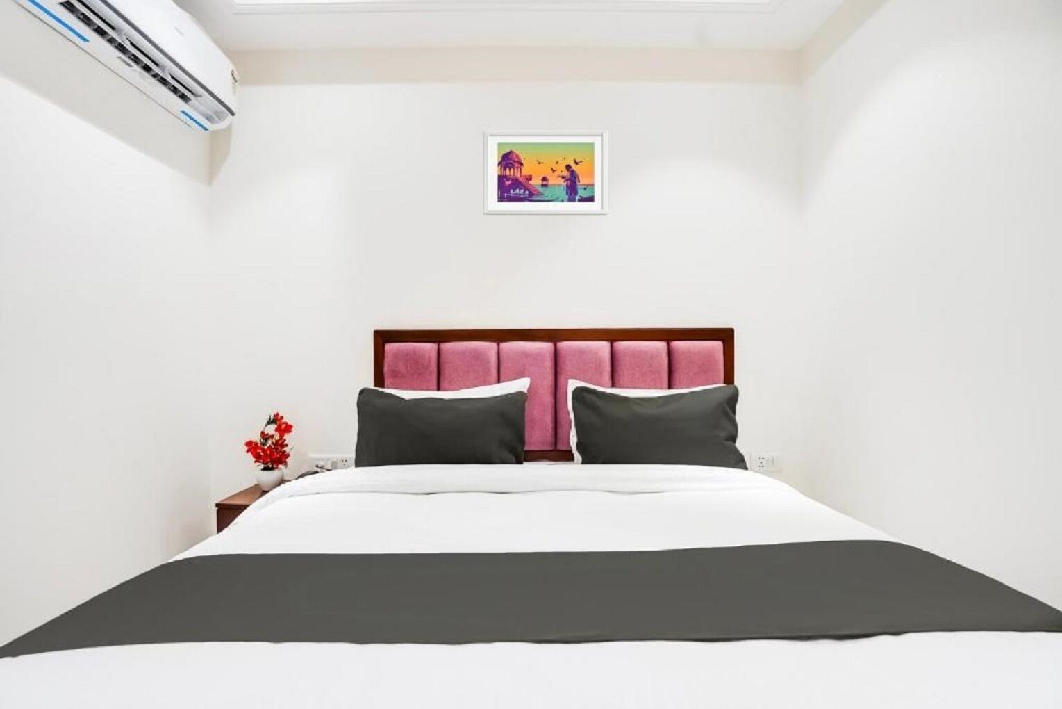 A bedroom with a bed, a nightstand, a vase with flowers, an air conditioning unit, and a framed picture on the wall.