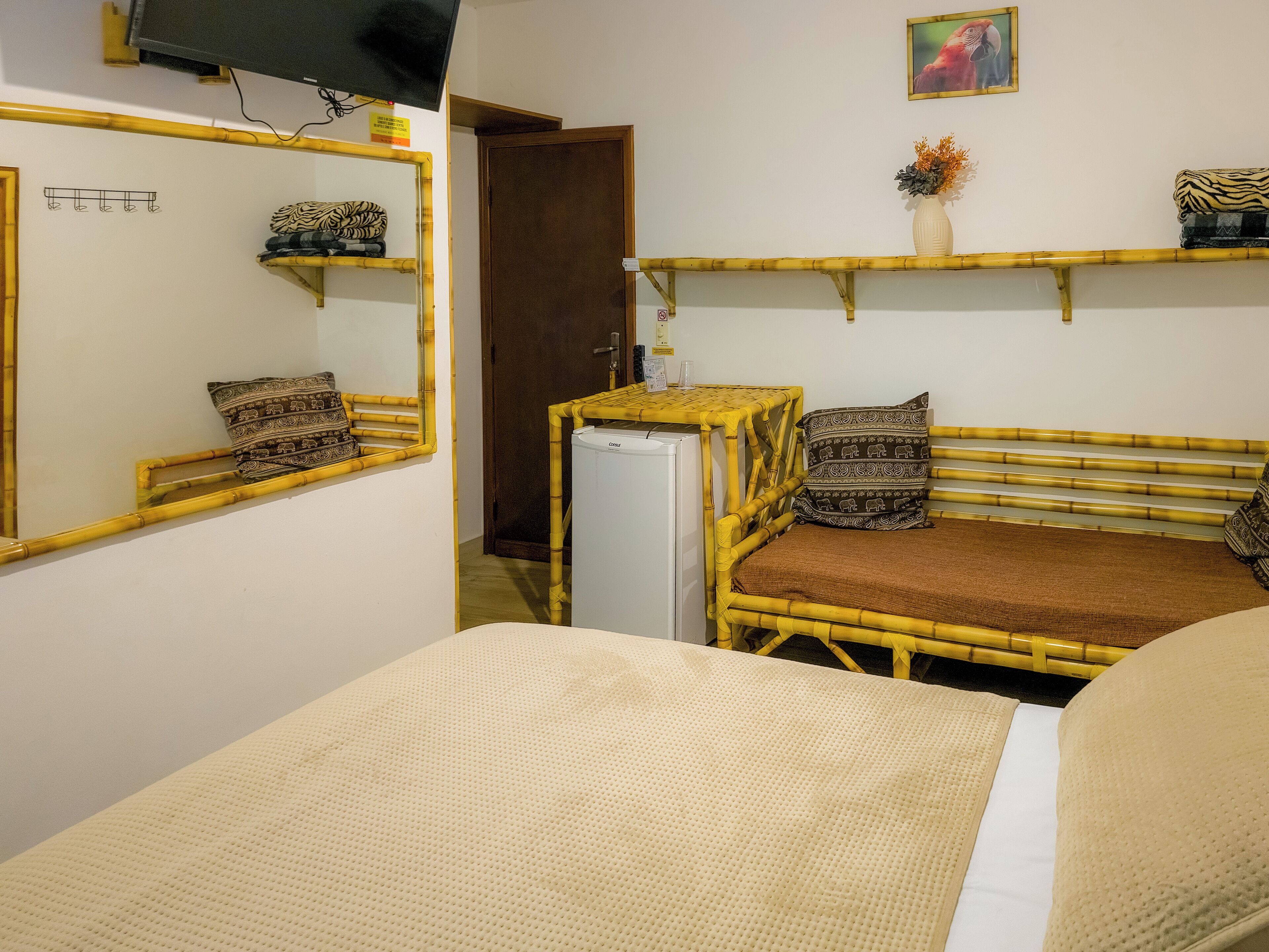 Una habitación con cama, una mesita, un refrigerador, un espejo y una estantería con adornos.