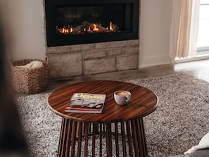 A round wooden table with a magazine and a cup, positioned in front of a stone fireplace with a visible fire. - Chalet Gratitude | Private SPA & Sauna | Skiing (Saint-Côme)