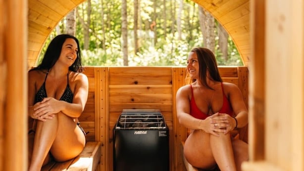 Deux personnes sont assises dans une cabane en bois, et un appareil de sauna portant l’inscription « HARVIA » est placé au centre.