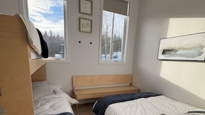 Une chambre avec deux lits superposés, une tête de lit en bois, une fenêtre donnant sur les arbres et un tableau encadré accroché au mur.