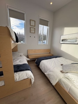 Une chambre avec deux lits superposés, une tête de lit en bois, une fenêtre donnant sur les arbres et un tableau encadré accroché au mur.