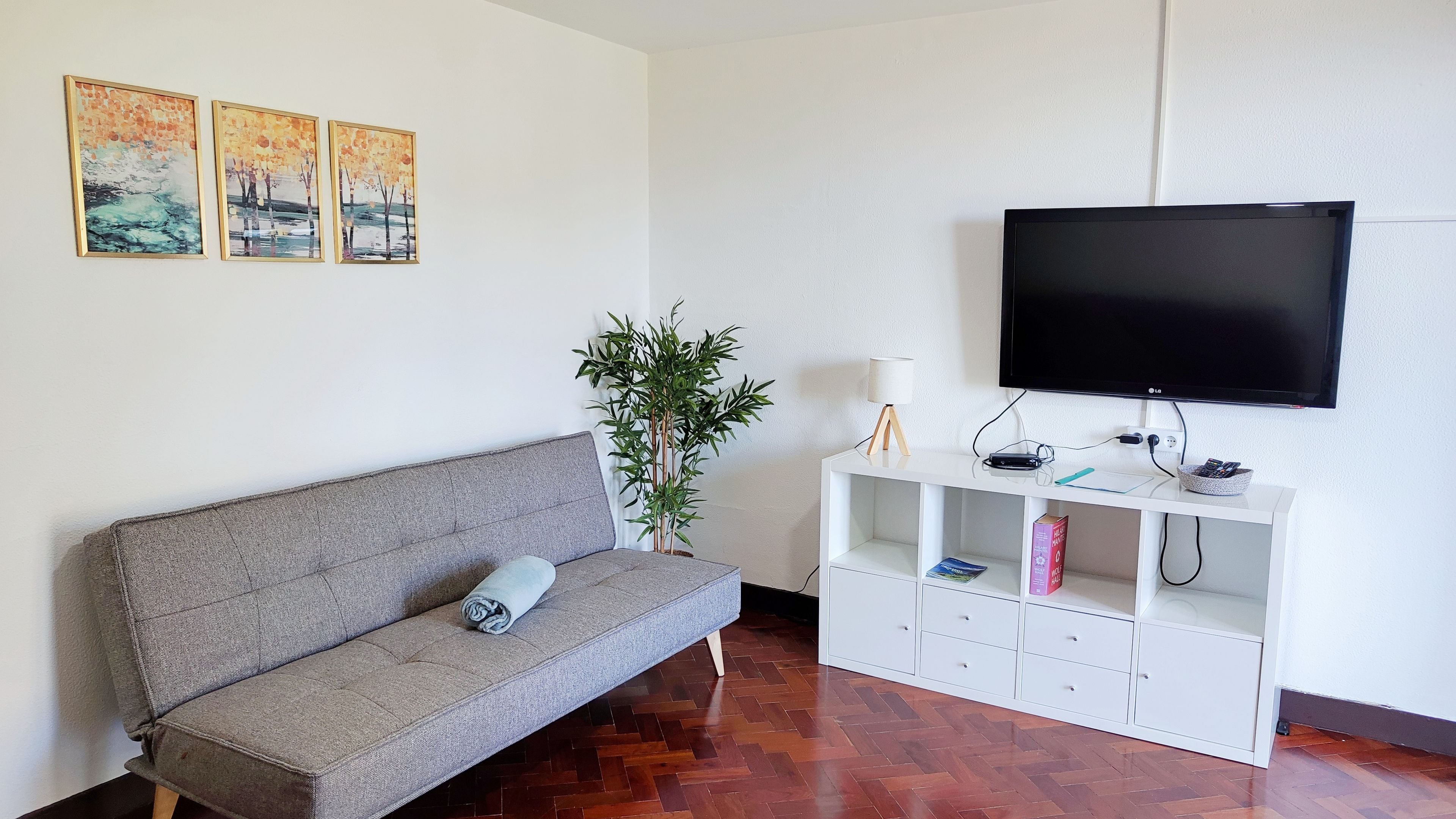 A living room with a grey sofa, a television mounted on the wall, a white entertainment unit with a lamp, and a potted plant.