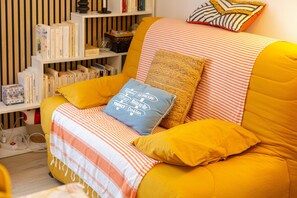 Un canapé jaune avec un couvre-lit à rayures et un coussin bleu orné de motifs, une bibliothèque remplie de livres et d’objets décoratifs, et un mur à rayures en arrière-plan.