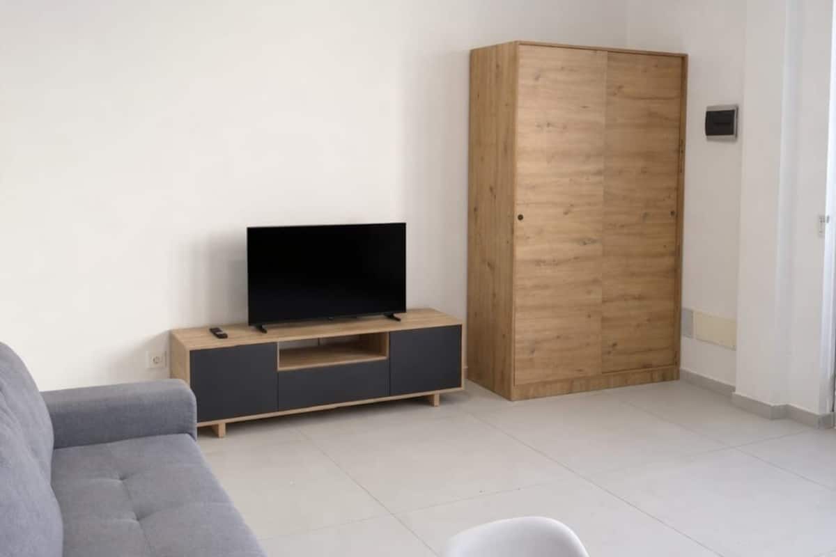A modern living room with a grey sofa, a wooden cabinet, a TV on a wooden stand, and a white chair.