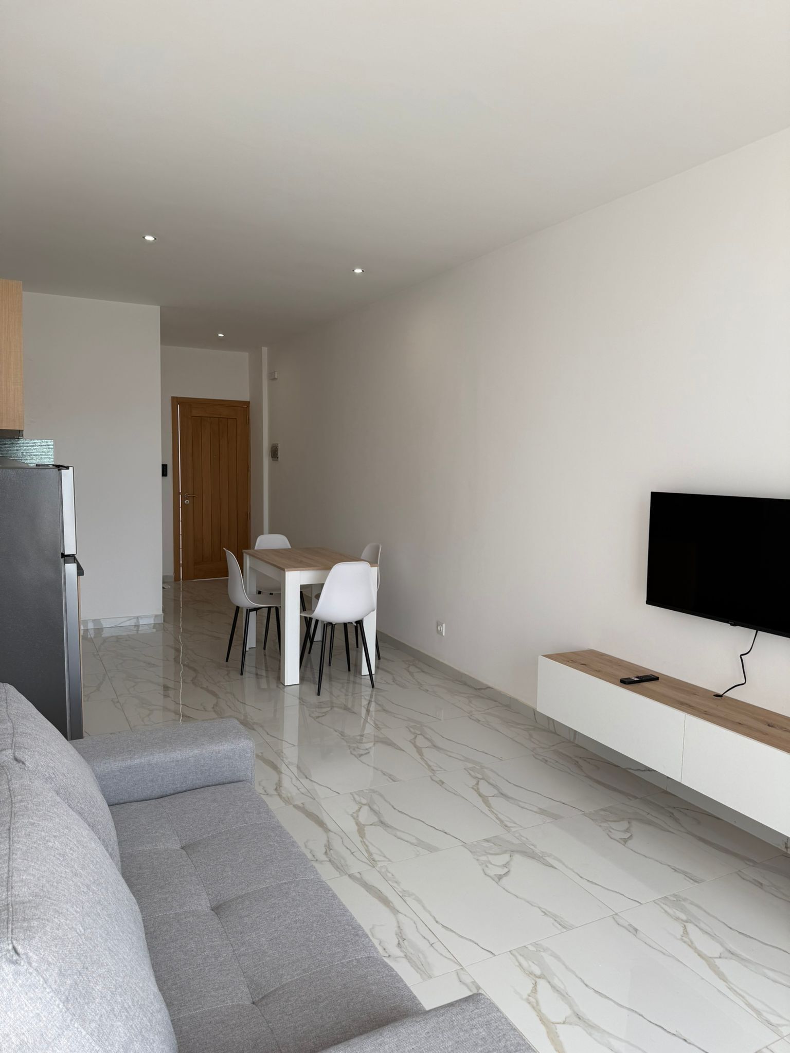 A modern living room with a grey sofa, a dining area with a wooden table and white chairs, a TV mounted on the wall, and a kitchen area with a fridge.