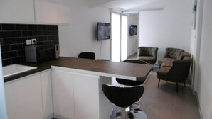 Een moderne keuken met witte kasten, een zwart betegelde spatwand, een houten aanrechtblad en een zwarte barkruk. Er is een zithoek met een bruine leren bank en twee fauteuils.