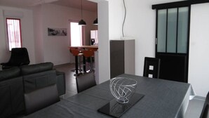 Een moderne woonkamer met een donkergrijze bank, een eettafel met een opvallend draadconstructie-decoratie, en een keuken in de achtergrond.
