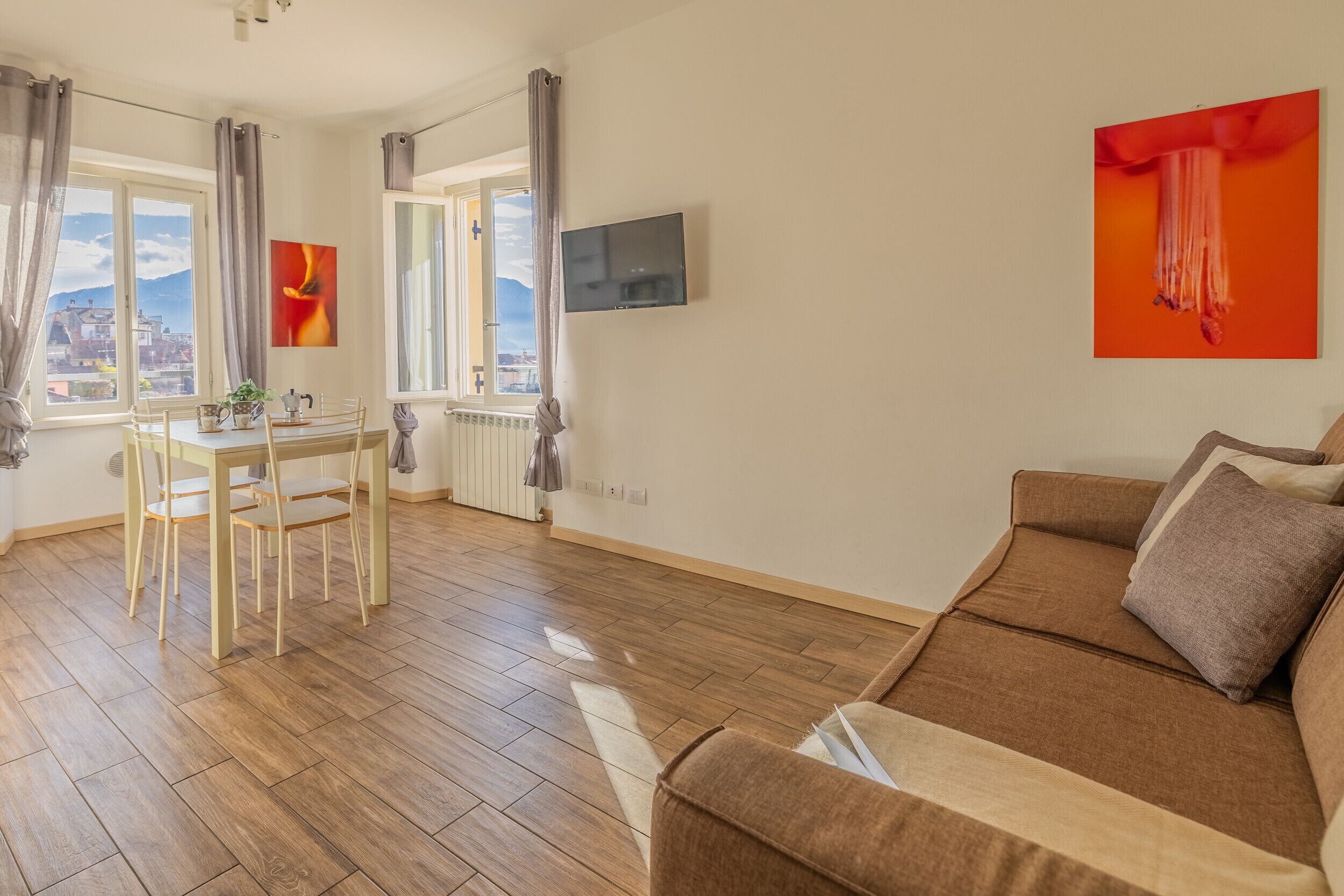 Een woonkamer met een bruine bank, een eettafel met stoelen, een aan de muur bevestigde televisie en een raam met gordijnen.