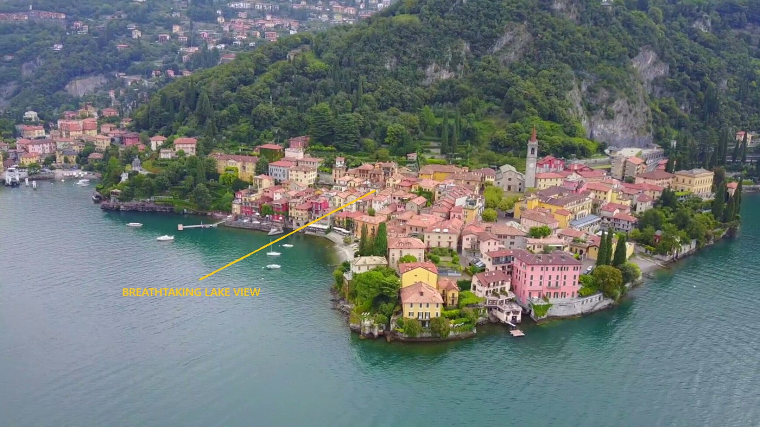 An aerial view of a lakeside town with buildings clustered around the water's edge, a prominent church tower, and a statue on a small island.