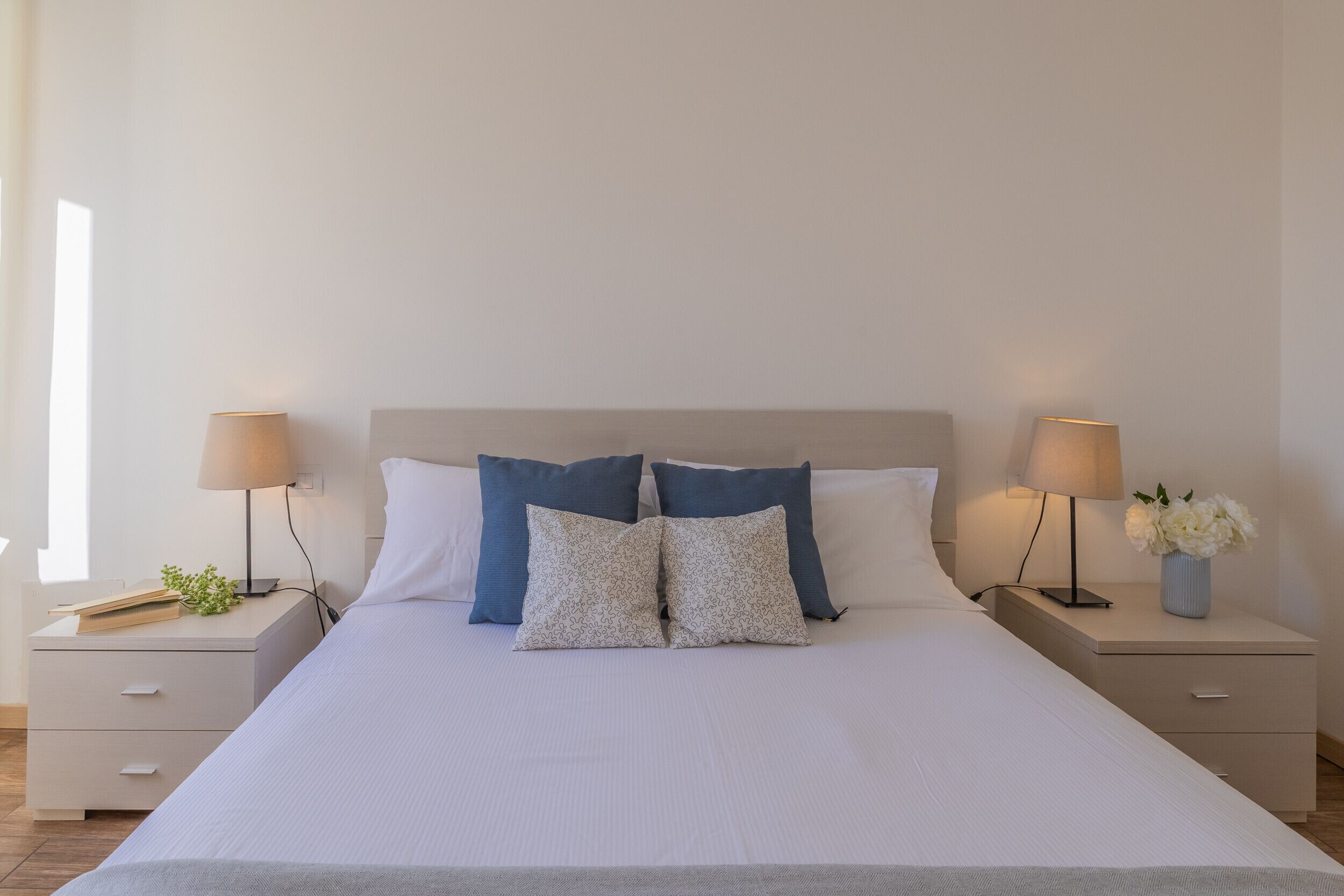 Un lit bien fait, avec du linge de lit blanc et deux coussins décoratifs, flanqué de deux tables de chevet sur lesquelles reposent des lampes et un petit vase de fleurs.