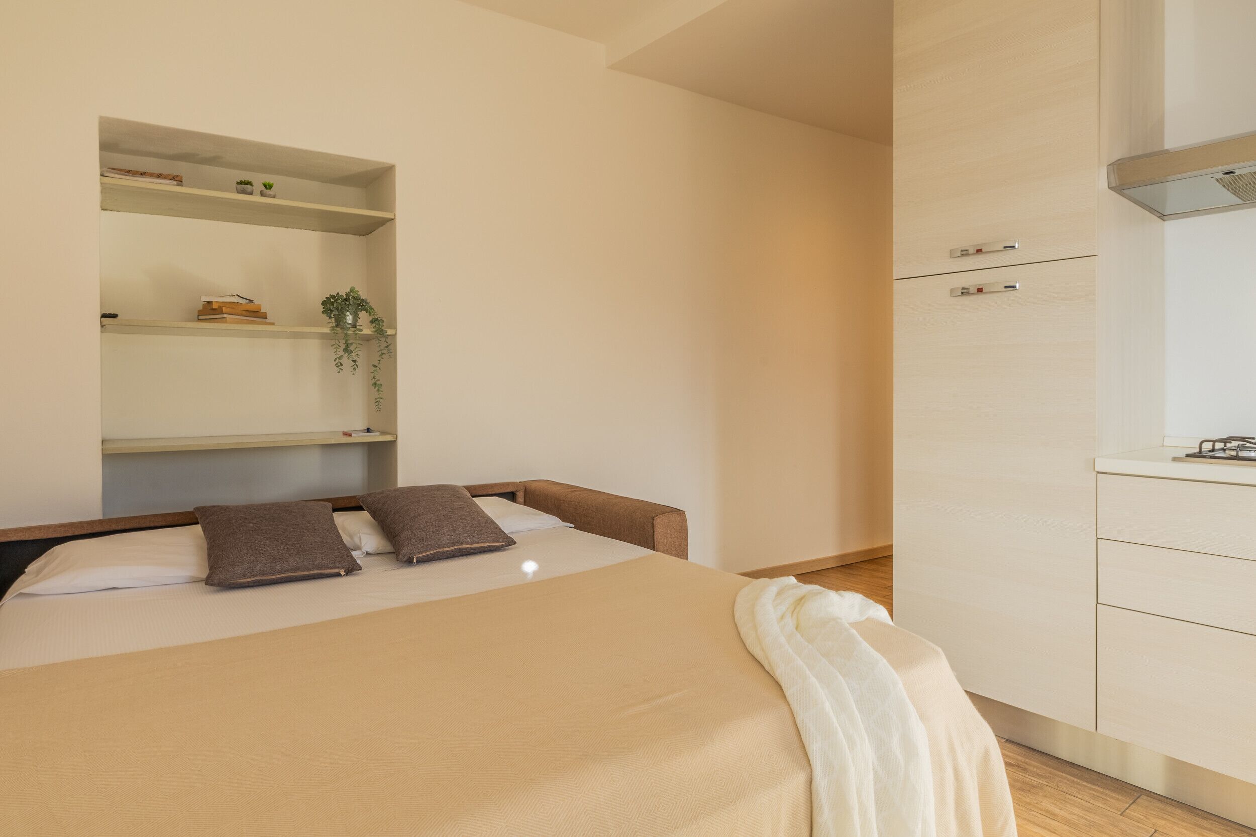 Un dormitorio con cama, una mesita de noche, una estantería con libros y una planta, además de una kitchenette con armarios y encimera.