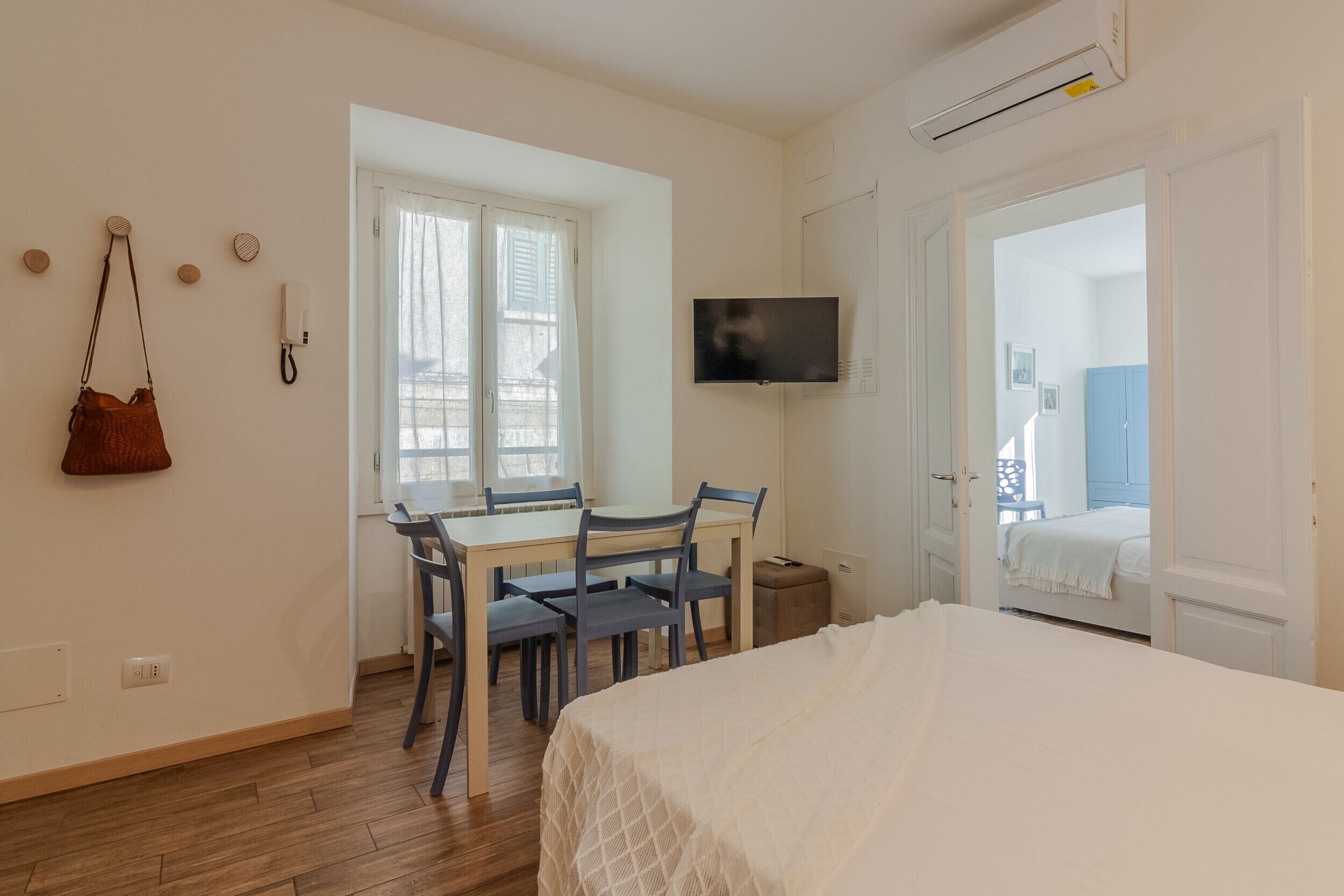 Ein Schlafzimmer mit Bett, einem an der Wand montierten Fernseher, einem Esstisch mit Stühlen und einem Fenster mit Vorhängen.