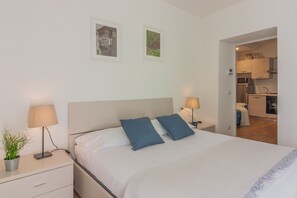 Une chambre à coucher avec un lit, deux oreillers, une table de chevet avec une lampe et une plante en pot. Une porte donne sur un espace cuisine.