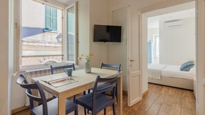 A room with a dining table, chairs, a TV mounted on the wall, a bed with a blue pillow, and a window with shutters. - Dream Lake Home in Varenna (Varenna)