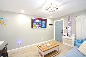 A living room with a flat-screen TV mounted on the wall, a wooden coffee table, a blue sofa, and a door with a sign above it. - Beautiful totally remodel condo with a pool and steps to the beach. (Ocean City)