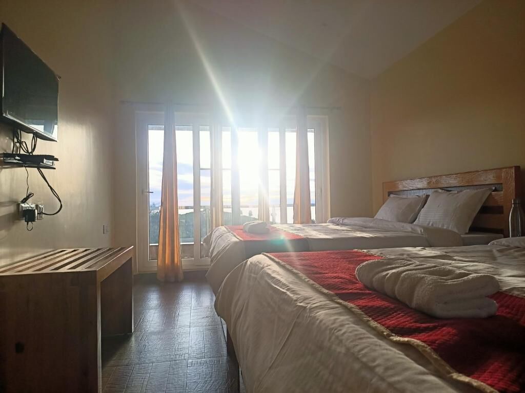 Camera d'albergo con due letti, una panca di legno, una TV a muro e un'ampia finestra con tende.