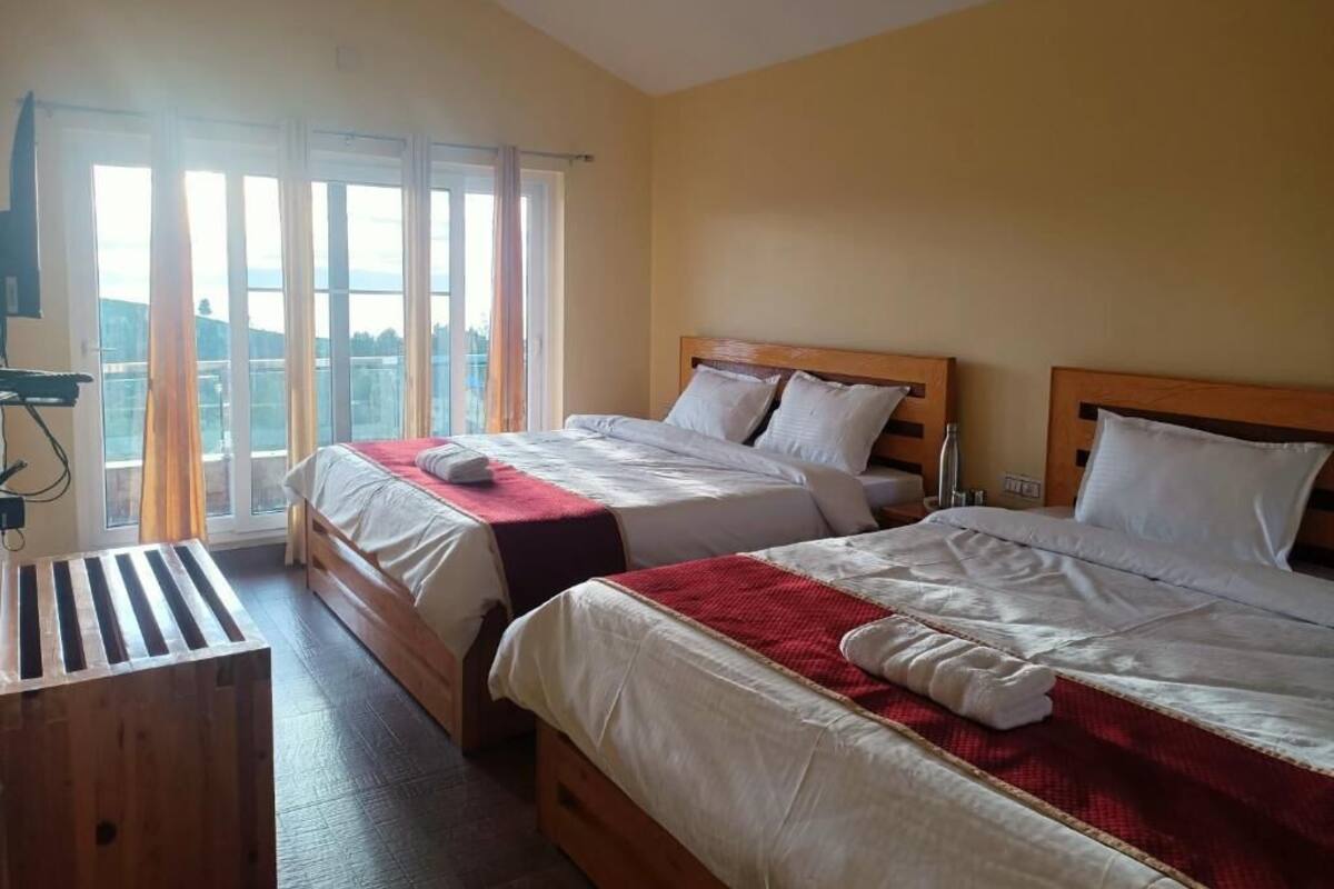A hotel room with two beds, a wooden bench, a window with curtains, and a view of the outside.