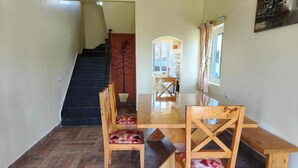 A dining area with a wooden table and chairs, a herringbone-patterned floor, a staircase, and a window with curtains. - Longwood Resorts (Kotagiri)