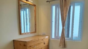A room with a wooden dresser, a mirror, a window with curtains, and a tiled floor. - Longwood Resorts (Kotagiri)