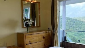 A hotel room with a bed, a wooden dresser, a mirror, and a window with a view of greenery. - Longwood Resorts (Kotagiri)