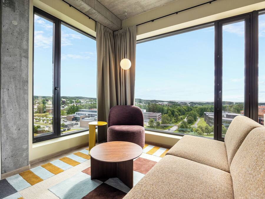 A modern hotel room with large windows, a sofa, a round table, and a chair.