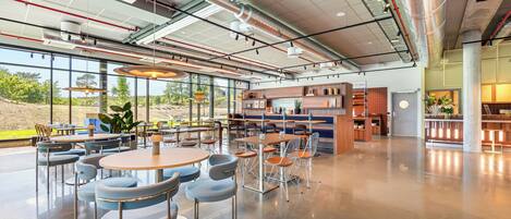 A spacious indoor area with tables and chairs, a kitchen area, and large windows offering a view of the outdoors.