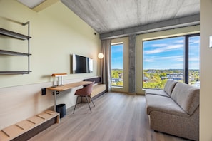 Ein modernes Zimmer mit einer Couch, einem Schreibtisch, einem Sessel, einem Fernseher und großen Fenstern mit Blick auf die Stadt.