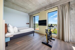 Moderni hotellihuone, jossa on sänky, kuntopyörä, suuret ikkunat ja televisio.