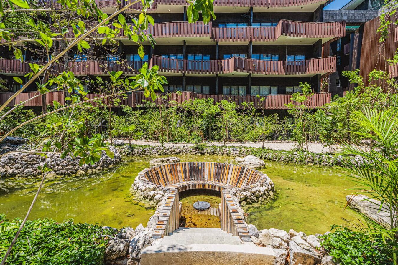 Un immeuble à plusieurs étages doté de balcons, entouré d’une végétation luxuriante et agrémenté d’une fontaine circulaire.