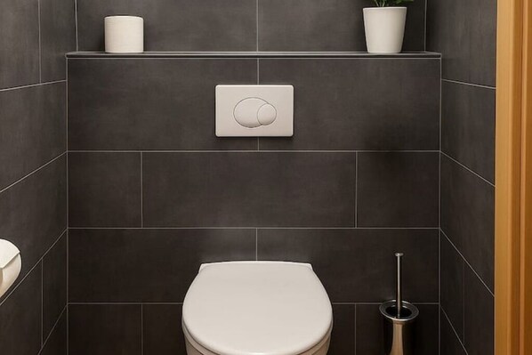A modern bathroom with dark tiles, a toilet, a bidet, a toilet paper holder, and a potted plant.