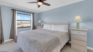 Quarto com cama, mesa de cabeceira, luminária, ventilador de teto e janela com cortinas.