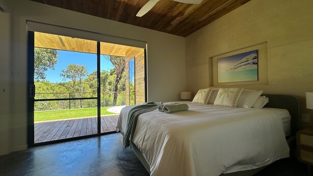A bedroom with a large bed, a painting on the wall, and a view of a green landscape through a sliding door. - Private coastal villa with traditional sauna located in a beautiful bush setting (Margaret River)