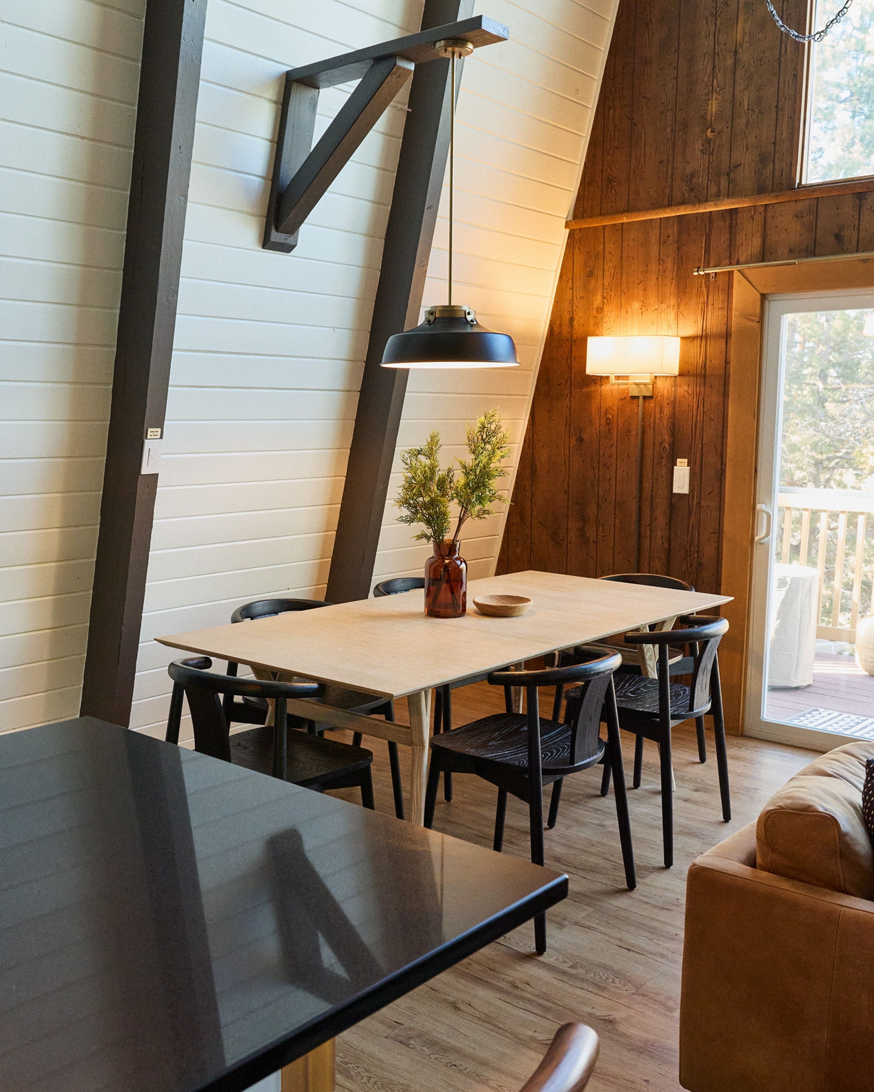 Un comedor con una mesa y sillas de madera, un florero con follaje e iluminación colgante.