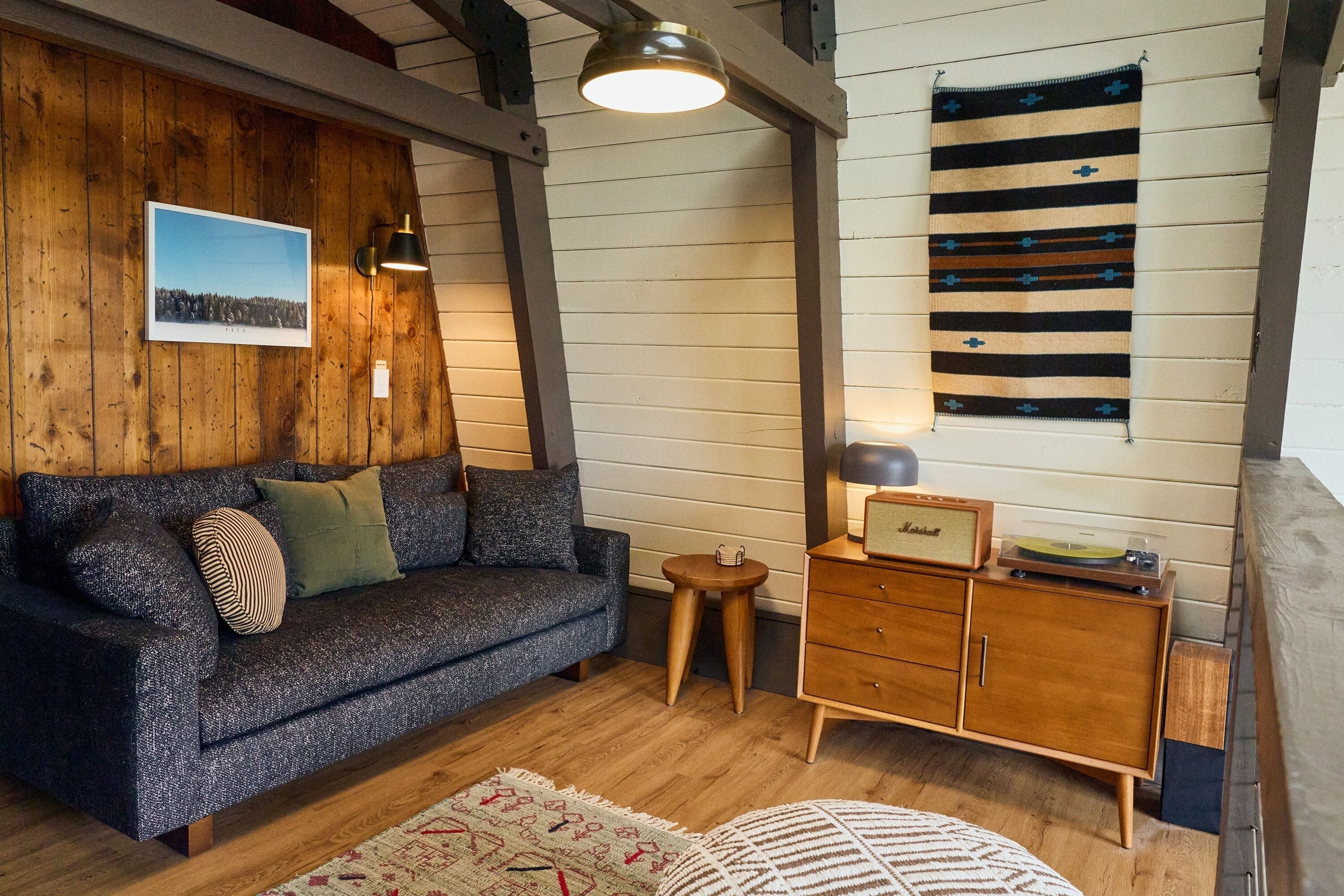 A cozy living room with a dark grey sofa, wooden walls, a small wooden cabinet, and a patterned rug.