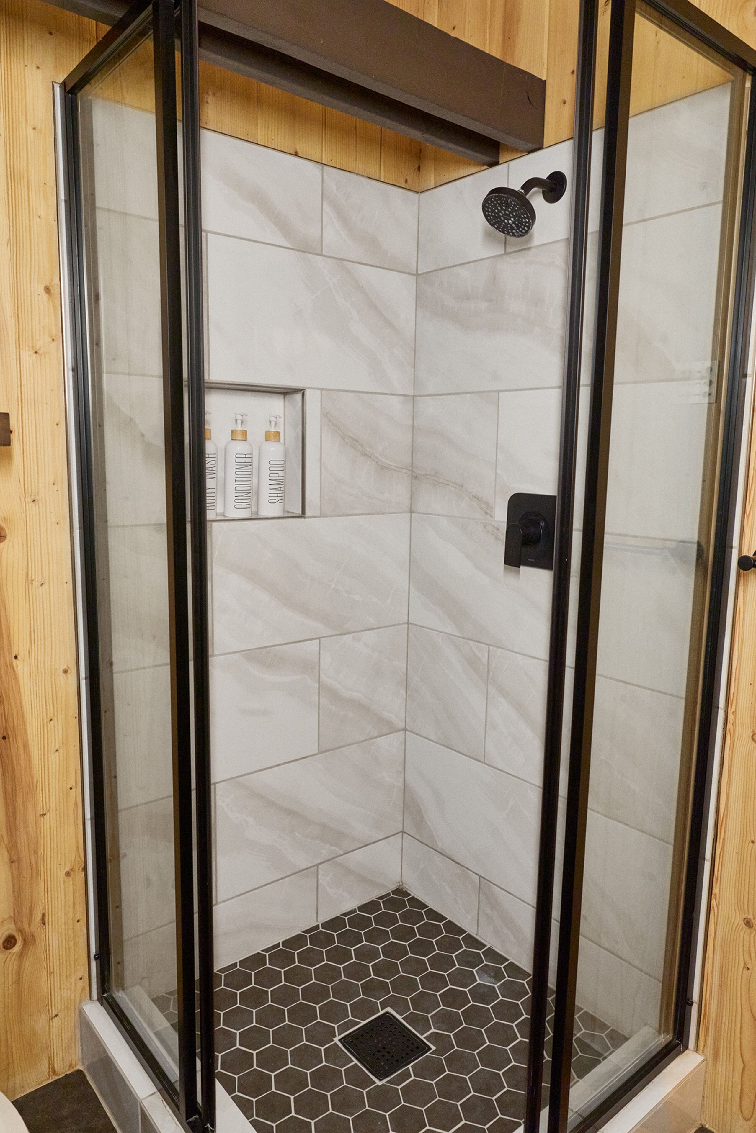 Una ducha en esquina con puertas de vidrio, azulejos de mármol blanco y un suelo con diseño hexagonal.