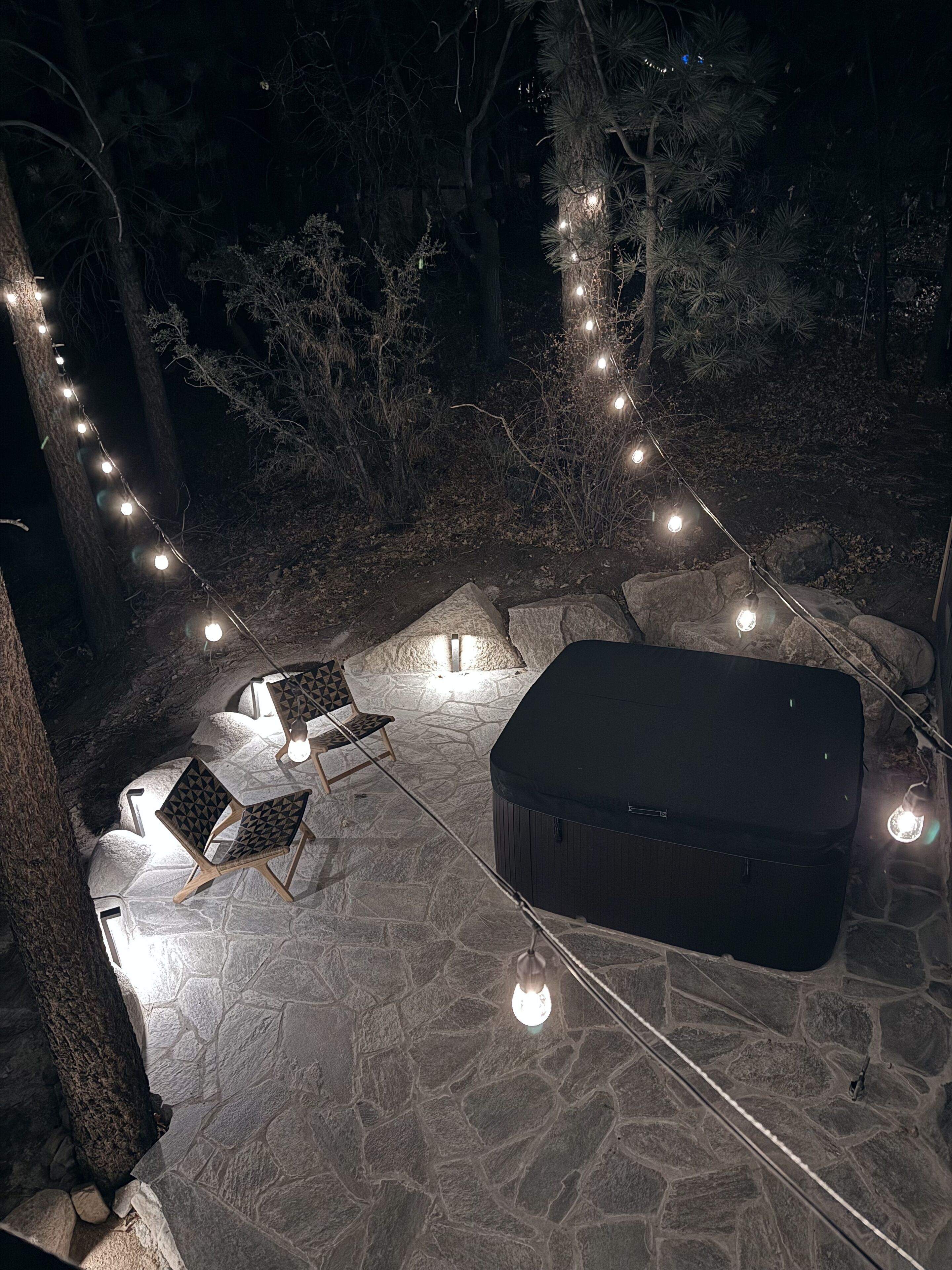 An outdoor patio with stone flooring, two lounge chairs, a hot tub, and string lights.