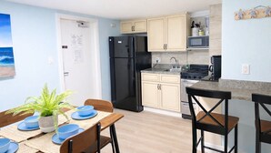 Eine kleine Küche mit Kühlschrank, Herd, Spüle und Holzausstattung. Ein Esstisch ist mit blauen Tellern und einem Topf mit Pflanze bestückt.